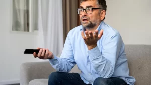 Homem surpreso segurando controle remoto enquanto a TV aparenta ter desligado sozinha.